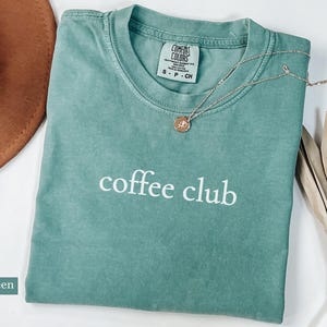 May include: Light green Comfort Colors t-shirt with the words "coffee club" in white. A gold necklace with a pendant rests on the shirt. A brown hat and dried plant are in the background.