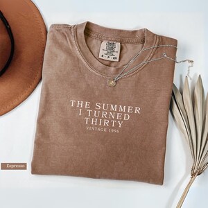 May include: Espresso-colored t-shirt with the text "THE SUMMER I TURNED THIRTY VINTAGE 1996" in white. A silver necklace with a pendant and a brown hat are also visible. The shirt is a Comfort Colors brand.