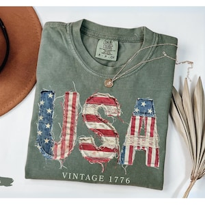 Puede incluir: Una camiseta verde musgo con la palabra "USA" aplicada en la parte delantera con un dise&ntilde;o de bandera estadounidense desgastada. La camiseta tambi&eacute;n presenta el texto "VINTAGE 1776". Un sombrero marr&oacute;n y un collar dorado tambi&eacute;n est&aacute;n en la imagen.