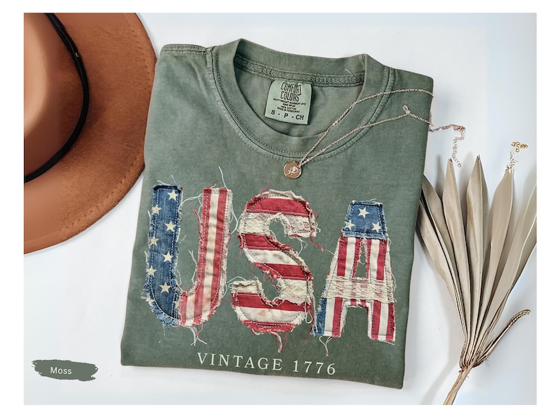 Puede incluir: Una camiseta verde musgo con la palabra "USA" aplicada en la parte delantera con un dise&ntilde;o de bandera estadounidense desgastada. La camiseta tambi&eacute;n presenta el texto "VINTAGE 1776". Un sombrero marr&oacute;n y un collar dorado tambi&eacute;n est&aacute;n en la imagen.