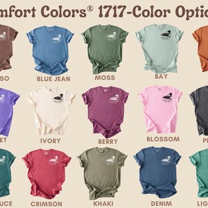 May include: A collection of Comfort Colors 1717 t-shirts in various colors, including Espresso, Blue Jean, Moss, and Yam. Each shirt features a small graphic on the chest and is displayed with its color name.