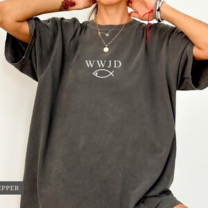 May include: Pepper-colored, oversized t-shirt featuring the text "WWJD" above a fish symbol in white. The shirt has short sleeves and a relaxed fit. The fabric appears soft and slightly faded.