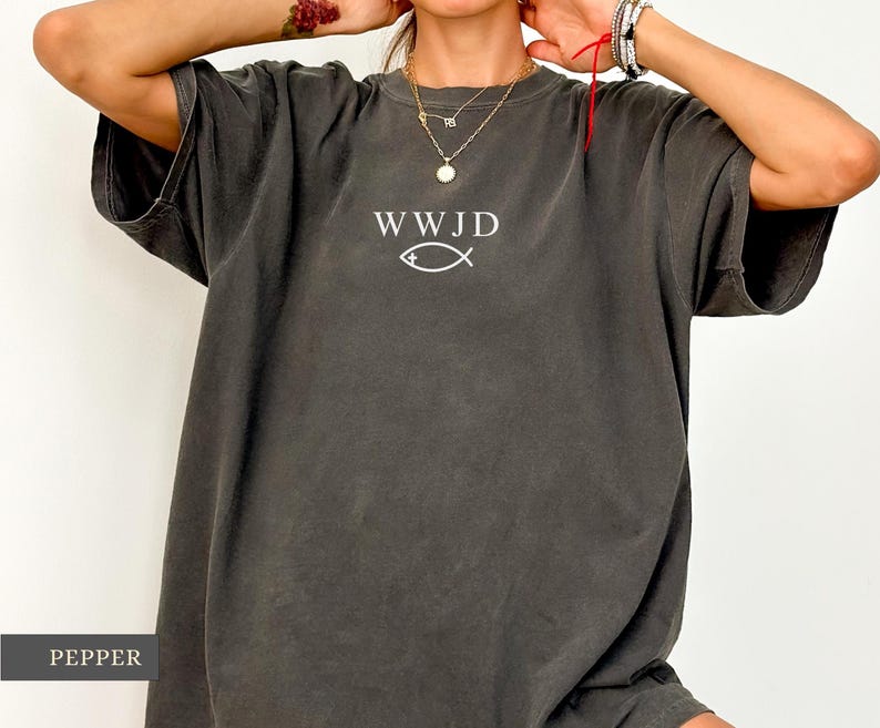 May include: Pepper-colored, oversized t-shirt featuring the text "WWJD" above a fish symbol in white. The shirt has short sleeves and a relaxed fit. The fabric appears soft and slightly faded.
