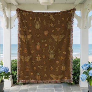 May include: A brown throw blanket with a repeating pattern of various insects, including beetles and moths, and floral accents. The blanket has a fringed edge and is displayed outdoors, hanging from a white structure with a view of the ocean.