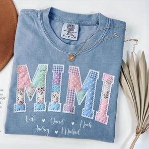 May include: Blue jean colored t-shirt with the word "MIMI" in a patchwork design. The shirt has the names "Kate", "David", "Audrey", "Noah", and "Michael" printed below the word "MIMI". A gold necklace and dried plant are also in the image.