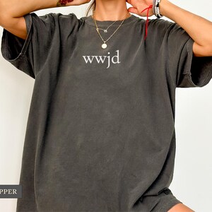Puede incluir: Camiseta de gran tama&ntilde;o color pimienta con el texto "wwjd" en blanco. La camiseta tiene mangas cortas y un ajuste relajado. La tela parece suave y c&oacute;moda, adecuada para uso casual.