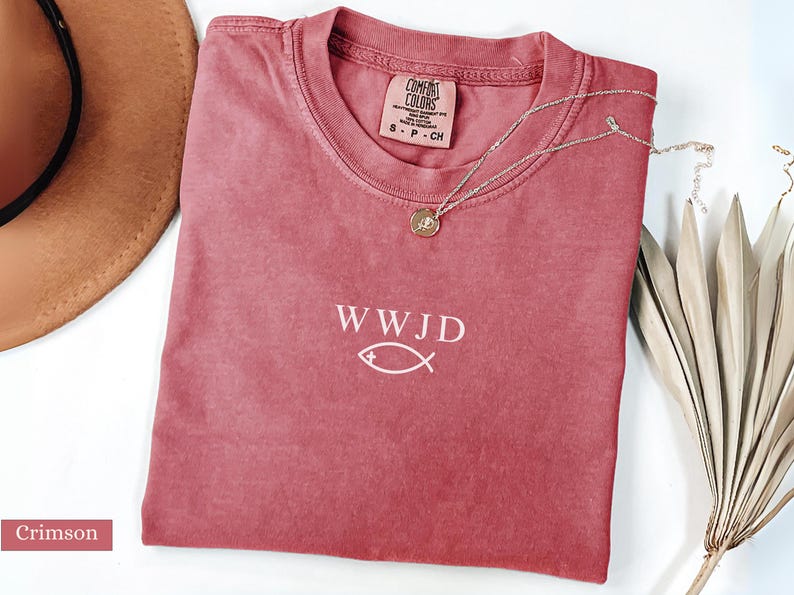 May include: Crimson-colored t-shirt with the text "WWJD" and a fish symbol. A silver necklace with a pendant rests on the shirt. The shirt is a Comfort Colors brand, size S-P-CH. A brown hat is visible in the background.