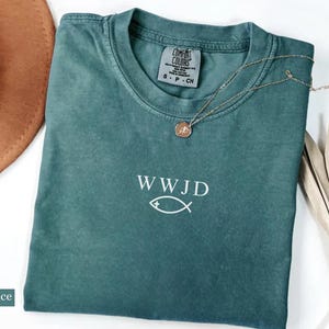 May include: Blue spruce t-shirt with the text "WWJD" and a fish symbol. A gold-colored necklace with a pendant rests on the shirt. A brown hat and dried plant are in the background. The shirt has a Comfort Colors tag.