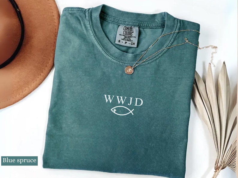 May include: Blue spruce t-shirt with the text "WWJD" and a fish symbol. A gold-colored necklace with a pendant rests on the shirt. A brown hat and dried plant are in the background. The shirt has a Comfort Colors tag.