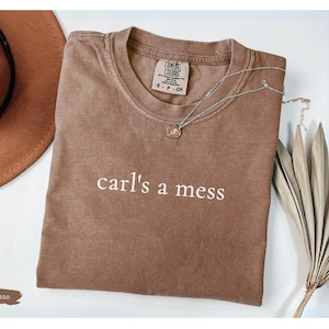 May include: Espresso-colored t-shirt with the text "carl's a mess" in white. The shirt is paired with a brown hat and a silver necklace. Dried decorative plants are also in the image.