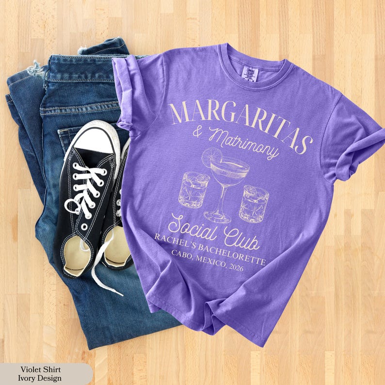 Może przedstawiać: Fioletowa koszulka z napisem "Margaritas & Matrimony Social Club" i grafiką szklanki do margarity. Koszulka jest połączona z niebieskimi dżinsami i czarnymi pł&oacute;ciennymi trampkami z białymi sznurowadłami. Widoczny jest r&oacute;wnież tekst "Rachel's Bachelorette Cabo, Mexico, 2026".