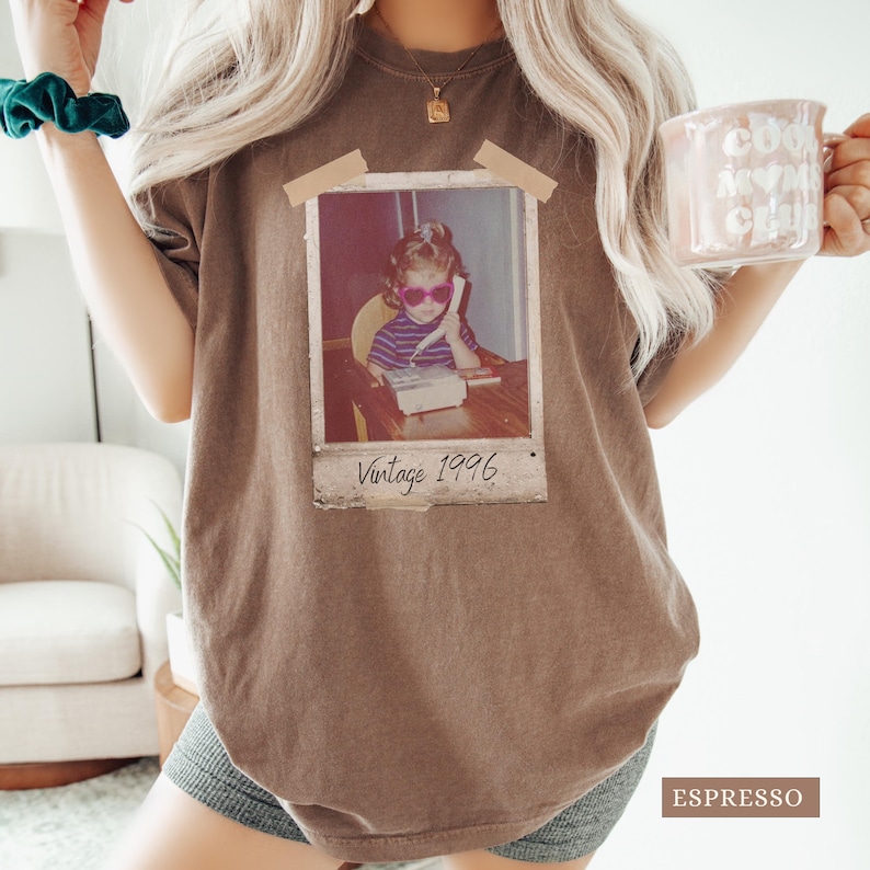 Pu&ograve; includere: T-shirt color espresso con un'immagine vintage in stile Polaroid di un bambino con occhiali da sole che usa un telefono. L'immagine &egrave; etichettata "Vintage 1996". La maglietta ha una vestibilit&agrave; ampia e maniche corte.