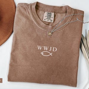 May include: Espresso-colored t-shirt with the text "WWJD" and a fish symbol. A silver necklace with a pendant rests on the shirt. A brown hat and dried plant stems are also visible.