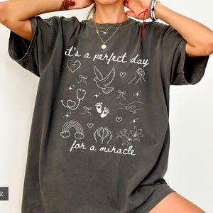 May include: A dark grey t-shirt with a white graphic design that says "it's a perfect day for a miracle". The graphic includes a dove, a stethoscope, a rainbow, and other symbols.