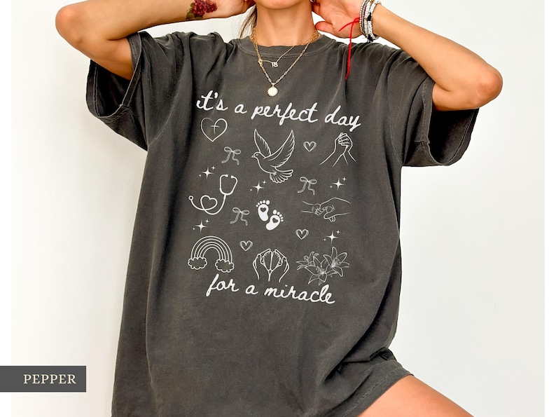 May include: A dark grey t-shirt with a white graphic design that says "it's a perfect day for a miracle". The graphic includes a dove, a stethoscope, a rainbow, and other symbols.