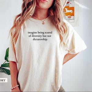 May include: Ivory-colored t-shirt with the text "imagine being scared of diversity but not dictatorship." The shirt has short sleeves and a relaxed fit. The word "IVORY" is printed at the bottom left.