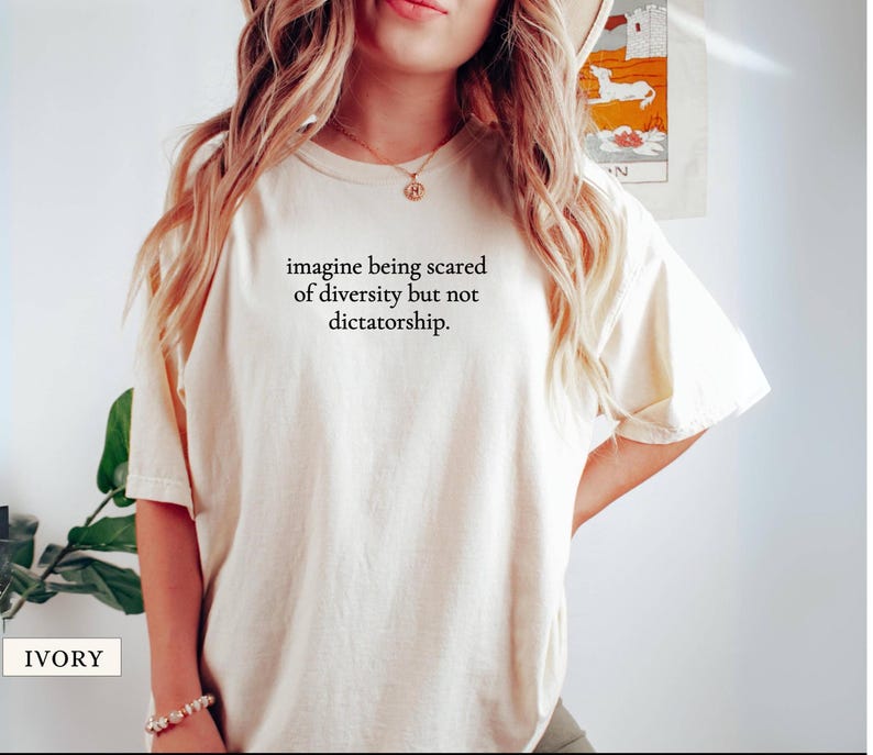 May include: Ivory-colored t-shirt with the text "imagine being scared of diversity but not dictatorship." The shirt has short sleeves and a relaxed fit. The word "IVORY" is printed at the bottom left.
