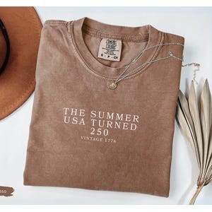 May include: Espresso-colored t-shirt with the text "THE SUMMER USA TURNED 250 VINTAGE 1776" in white. A brown hat and a silver necklace with a pendant are also visible. The shirt is a Comfort Colors brand.