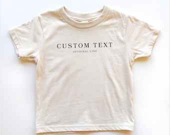 Może przedstawiać: Kremowy t-shirt z krótkim rękawem z czarnym nadrukiem na klatce piersiowej. Napis brzmi CUSTOM TEXT dużą czcionką, a słowa OPTIONAL LINE są wydrukowane mniejszą czcionką bezpośrednio pod główną linią tekstu.