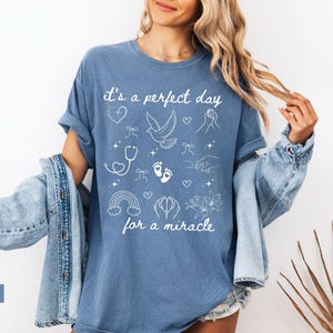 May include: A blue jean t-shirt with white text that reads "It's a perfect day for a miracle". The shirt is decorated with white illustrations of a dove, a rainbow, a stethoscope, a heart, and footprints.
