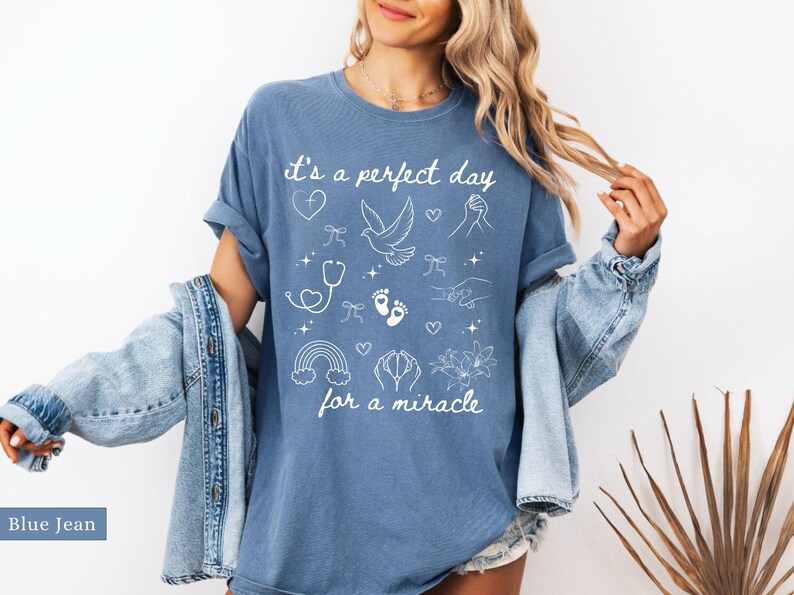 May include: A blue jean t-shirt with white text that reads "It's a perfect day for a miracle". The shirt is decorated with white illustrations of a dove, a rainbow, a stethoscope, a heart, and footprints.