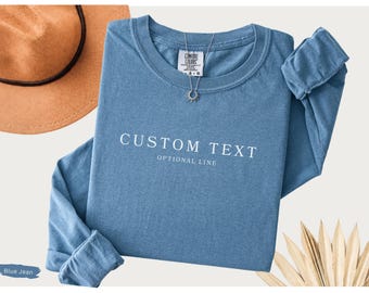 May include: A blue long-sleeve shirt with a crew neckline and a Comfort Colors label at the inner collar. The front features the white text CUSTOM TEXT and OPTIONAL LINE. A silver sun-shaped pendant necklace rests on the shirt fabric.