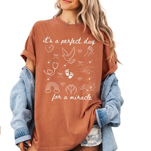 May include: A rust-coloured t-shirt with a white graphic design. The graphic features a dove, hearts, footprints, a stethoscope, a rainbow, and the text "It's a perfect day for a miracle".