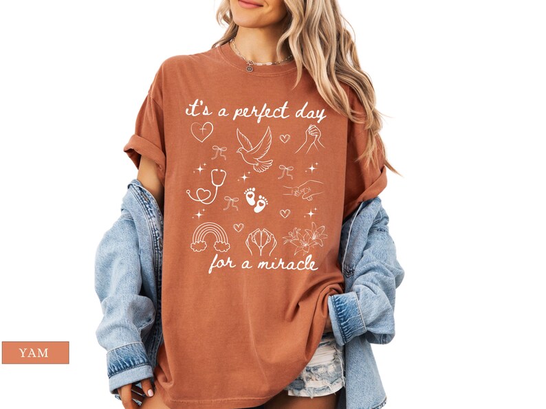 May include: A rust-coloured t-shirt with a white graphic design. The graphic features a dove, hearts, footprints, a stethoscope, a rainbow, and the text "It's a perfect day for a miracle".