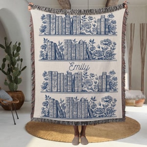 May include: Cream-colored throw blanket featuring a blue bookshelf design with floral accents. The blanket has three shelves of books and the name "Emily" in the center. The edges are fringed.