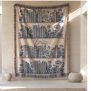 May include: A beige throw blanket with a blue bookshelf design. The design includes rows of books, floral accents, and the name "Emily" in script. The blanket has a fringed edge and is displayed on a wall.