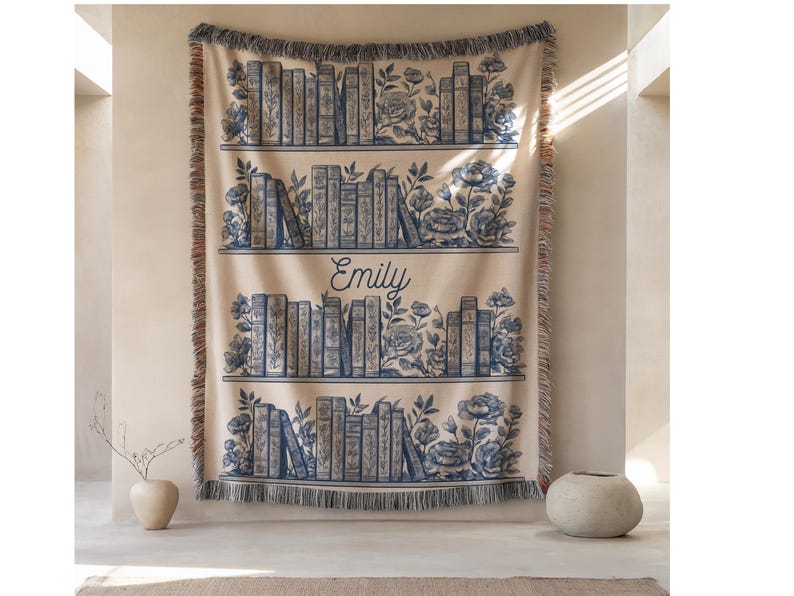 May include: A beige throw blanket with a blue bookshelf design. The design includes rows of books, floral accents, and the name "Emily" in script. The blanket has a fringed edge and is displayed on a wall.