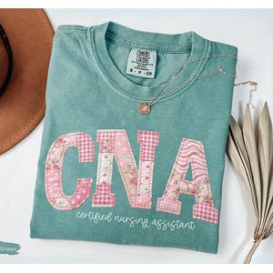 May include: Light green t-shirt featuring the letters "CNA" in pink floral and gingham fabric. The words "certified nursing assistant" are printed below the letters in white. A gold necklace and a brown hat are also visible.