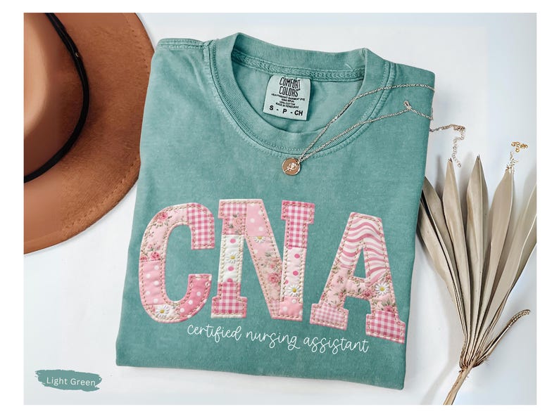 May include: Light green t-shirt featuring the letters "CNA" in pink floral and gingham fabric. The words "certified nursing assistant" are printed below the letters in white. A gold necklace and a brown hat are also visible.