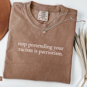 May include: Espresso-colored t-shirt with the text "stop pretending your racism is patriotism." A silver necklace with a small pendant rests on the shirt. The shirt is folded on a white surface, with a brown hat and dried decorative plants in the background.