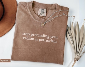May include: Espresso-colored t-shirt with the text "stop pretending your racism is patriotism." A silver necklace with a small pendant rests on the shirt. The shirt is folded on a white surface, with a brown hat and dried decorative plants in the background.