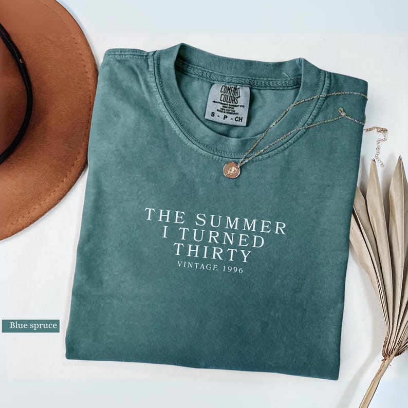 May include: A blue spruce Comfort Colors t-shirt with the text "THE SUMMER I TURNED THIRTY VINTAGE 1996" in white. A gold necklace with a pendant and a brown hat are also in the image.