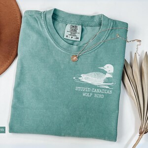 May include: Light green Comfort Colors t-shirt with a white graphic of a loon and the text "STUPID CANADIAN WOLF BIRD". A gold necklace and a brown hat are also in the image. The shirt is size S-P-CH.