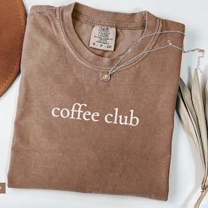 May include: Espresso-coloured t-shirt with the words "coffee club" in white. A silver necklace with a small pendant rests on the shirt. The shirt is folded on a white surface, with a brown hat and dried plants in the background.