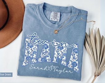 May include: A blue jean t-shirt with the words "twin mama" in a blue and white floral print. The names "Sarah" and "Taylor" are written below the words "twin mama" with a heart symbol between the names.