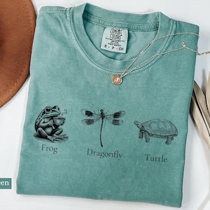 May include: Light green t-shirt with a frog, dragonfly, and turtle graphic. The shirt has the text "Comfort Colors" and "S - P - CH" printed on the tag. The text below the graphic reads "Frog", "Dragonfly", and "Turtle".