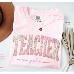 May include: A light pink t-shirt with the word "TEACHER" in a patchwork design, featuring floral and gingham patterns. The name "mrs. johnson" is written below. A necklace and a brown hat are also visible.