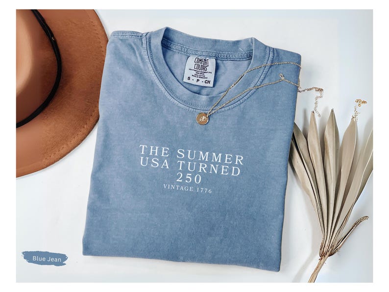 May include: A blue jean colored t-shirt with the text "THE SUMMER USA TURNED 250 VINTAGE 1776" in white. A gold necklace and a brown hat are also in the image. The shirt is a Comfort Colors brand.