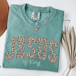 Op de afbeelding: Lichtgroen T-shirt met de tekst "JESUS is King" in een luipaardprint. Het shirt heeft een ronde hals en korte mouwen. Een gouden ketting met een hanger is over het shirt gedrapeerd.
