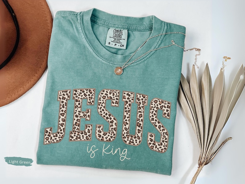 Op de afbeelding: Lichtgroen T-shirt met de tekst "JESUS is King" in een luipaardprint. Het shirt heeft een ronde hals en korte mouwen. Een gouden ketting met een hanger is over het shirt gedrapeerd.