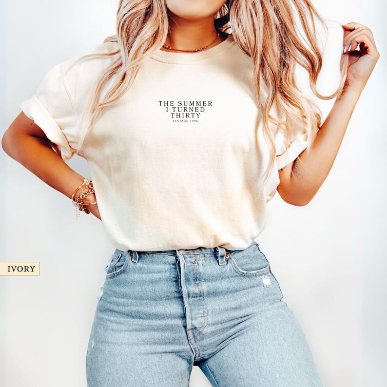 May include: Ivory-colored t-shirt with the text "THE SUMMER I TURNED THIRTY VINTAGE 1994" in black. The shirt is paired with light blue jeans. The word "IVORY" is in a box in the lower left corner.