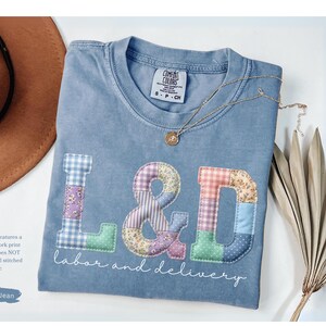 May include: A blue jean Comfort Colors t-shirt with a faux patchwork print of the letters "L&D" and the words "labor and delivery". The letters are in various colors and patterns. A gold necklace and a brown hat are also in the image.