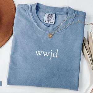 Puede incluir: Una camiseta azul vaquero Comfort Colors con el texto "wwjd" en blanco. Un collar de oro con un colgante descansa sobre la camisa. Un sombrero marr&oacute;n y una planta seca est&aacute;n en el fondo.