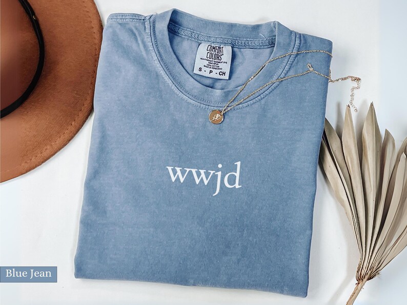 Puede incluir: Una camiseta azul vaquero Comfort Colors con el texto "wwjd" en blanco. Un collar de oro con un colgante descansa sobre la camisa. Un sombrero marr&oacute;n y una planta seca est&aacute;n en el fondo.