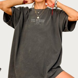 May include: Dark gray oversized t-shirt with short sleeves. The shirt features the text "THE SUMMER USA TURNED 250 VINTAGE 1776" in a small font on the front. The shirt has a vintage, worn look.