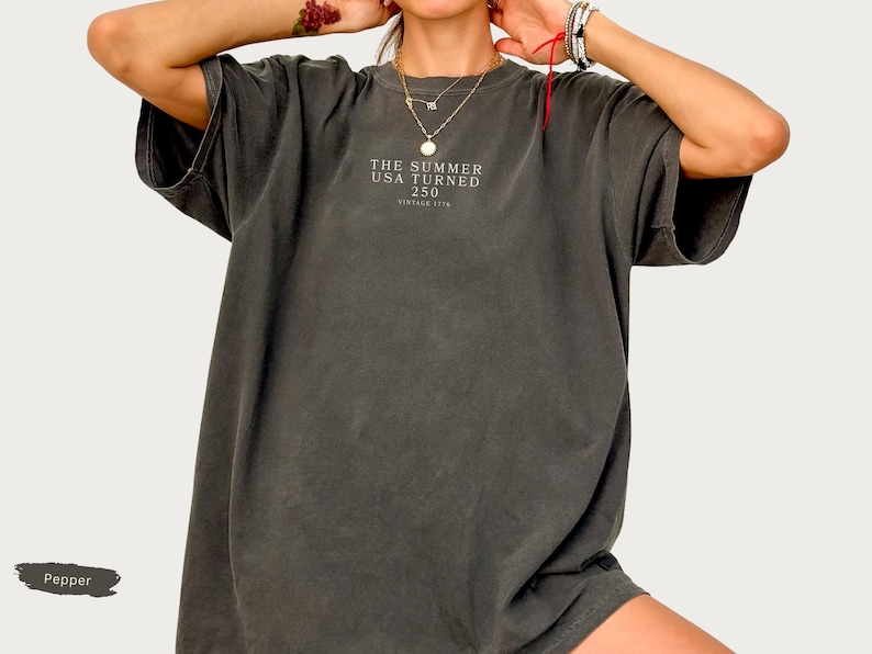 May include: Dark gray oversized t-shirt with short sleeves. The shirt features the text "THE SUMMER USA TURNED 250 VINTAGE 1776" in a small font on the front. The shirt has a vintage, worn look.